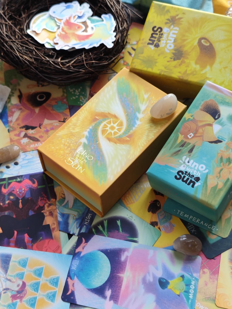 May include: An assortment of tarot cards and boxes with the text "Uno with the Sun". The boxes are yellow, orange, and teal. The cards feature colorful illustrations and are scattered around a small bird's nest with stickers.