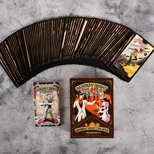 May include: A deck of tarot cards with black card backs and gold edges, fanned out on a gray surface. The box and card backs feature the text "Fantasy Night Theater" and illustrations of figures in vintage attire. One card shows "The Fool".