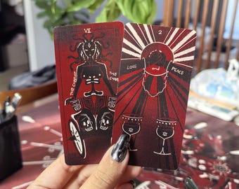 Puede incluir: Dos cartas de tarot con diseños rojos y negros se sostienen. Una carta presenta un carro con una figura y el texto "THE CHARIOT". La otra carta tiene las palabras "LOVE and PEACE" y dos cálices.