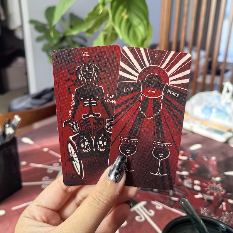 Puede incluir: Dos cartas de tarot con dise&ntilde;os rojos y negros se sostienen. Una carta presenta un carro con una figura y el texto "THE CHARIOT". La otra carta tiene las palabras "LOVE and PEACE" y dos c&aacute;lices.