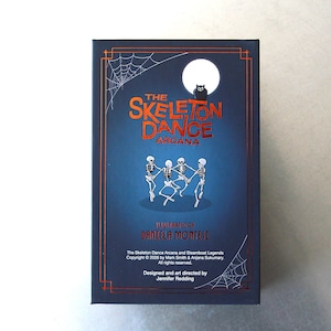 May include: A dark blue box featuring the text "The Skeleton Dance Arcana" in orange. The image includes illustrations of dancing skeletons, a full moon, a black owl, and spiderwebs. The box is designed by Jennifer Redding and illustrated by Daniela Montesi.
