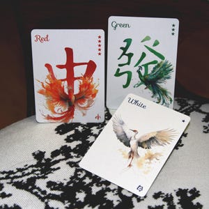 Pu&ograve; includere: Tre carte da gioco con illustrazioni ad acquerello. La carta rossa presenta un disegno di fenice e caratteri cinesi. La carta verde ha caratteri cinesi e un pavone. La carta bianca mostra una gru. Le carte sono su una superficie a motivi.