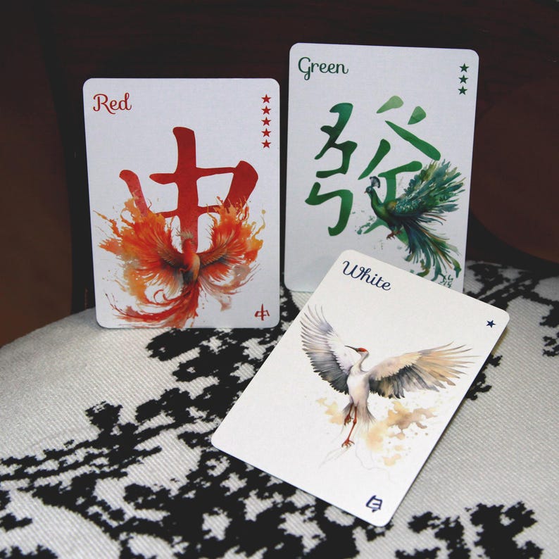Pu&ograve; includere: Tre carte da gioco con illustrazioni ad acquerello. La carta rossa presenta un disegno di fenice e caratteri cinesi. La carta verde ha caratteri cinesi e un pavone. La carta bianca mostra una gru. Le carte sono su una superficie a motivi.