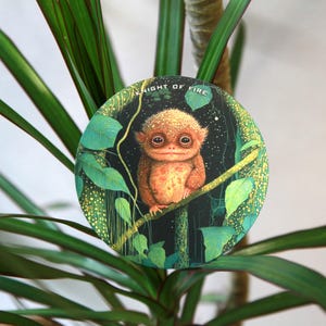 May include: A round illustration featuring a creature with large eyes and orange fur, perched on a branch. The background is dark with green leaves and the text "KNIGHT OF FIRE". The image is set against a backdrop of green plant leaves.