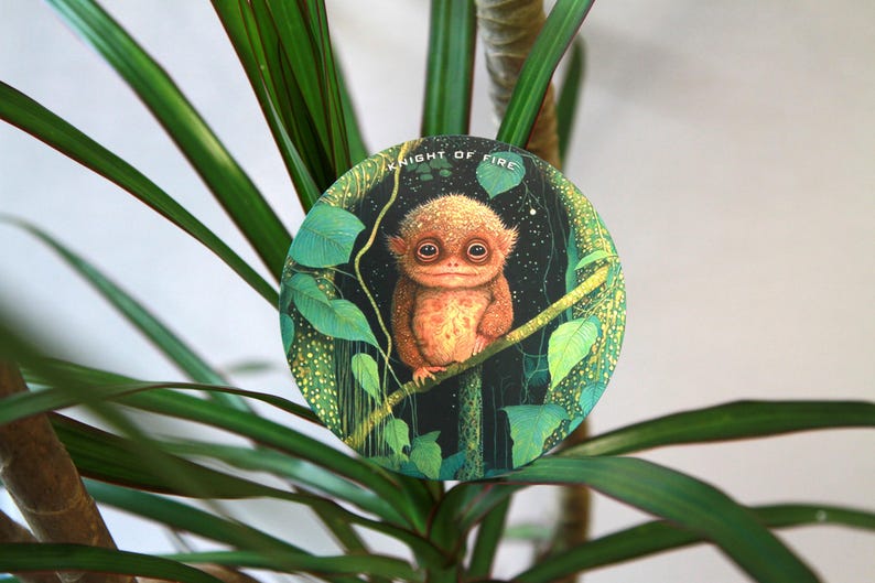 May include: A round illustration featuring a creature with large eyes and orange fur, perched on a branch. The background is dark with green leaves and the text "KNIGHT OF FIRE". The image is set against a backdrop of green plant leaves.