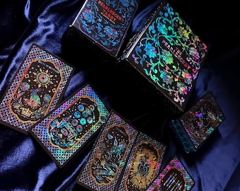 Könnte beinhalten: Ein Pearlescent Tarot-Kartendeck mit schwarzem Hintergrund und irisierenden, holografischen Folienillustrationen von Blumenmustern, himmlischen Symbolen und Fabelwesen. Das Set enthält eine große Schachtel und mehrere einzelne Karten.