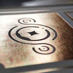 May include: A gold metallic background with a black design of a circle with a diamond in the center and two smaller circles around it.