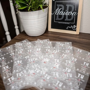 Puede incluir: Primer plano de una pila de etiquetas de ropa de plástico transparente con el texto "T.U. Made in USA" impreso en ellas. Las etiquetas están dispuestas de forma aleatoria sobre una superficie de madera.