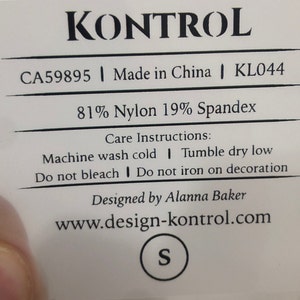 Puede incluir: Una etiqueta blanca con texto negro que dice "KONTROL", "CA59895 | Made in China | KL044", "81% Nylon 19% Spandex", "Instrucciones de cuidado: Lavar a máquina con agua fría | Secar en secadora a baja temperatura | No usar lejía | No planchar sobre la decoración", "Diseñado por Alanna Baker", "www.design-kontrol.com" y "S".