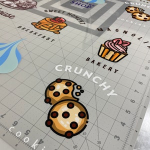 May include: A set of colorful stickers featuring various bakery items, including cupcakes, cookies, and pancakes. The stickers are arranged on a gray grid background with the words "Sweet Moment", "Cupcake Shop", "Bakery", "Crunchy", and "Magnolia" visible.