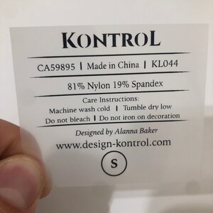 Puede incluir: Una etiqueta de ropa blanca con texto negro. La etiqueta dice "KONTROL", "CA59895 | Made in China | KL044", "81% Nylon 19% Spandex", "Instrucciones de cuidado: Lavar a máquina con agua fría | Secar en secadora a baja temperatura", "No usar lejía | No planchar sobre la decoración", "Diseñado por Alanna Baker", "www.design-kontrol.com", y "S".