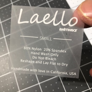 Puede incluir: Una etiqueta blanca con el texto "Laello swimwear" y "SMALL" impreso en ella. La etiqueta también incluye instrucciones de cuidado: "80% Nylon 20% Spandex, Lavar a mano únicamente, No usar lejía, Dar forma y dejar secar plano". La etiqueta concluye con "Hecho a mano con amor en California, EE. UU.".