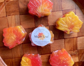 May include: Five maple leaf-shaped soaps in shades of orange and red, wrapped in clear plastic. The soaps are arranged on a wooden tray. The label reads "Maple Leaf, maple syrup scented honey soap, handcrafted in Mackinaw, MI, net wt 2.3 oz / 70 g."