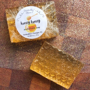 May include: Two bars of honey scented soap, one wrapped in clear plastic with a label that reads "honey honey scented honey soap handcrafted in Cheboygan, MI Net Wt 4 oz | 113 g", the other bar is unwrapped and has a honeycomb pattern.