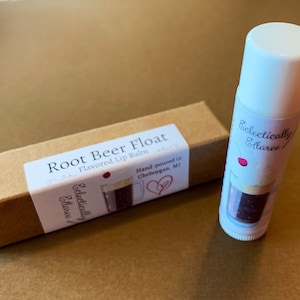 May include: A brown cardboard box with a white label that reads "Root Beer Float Flavored Lip Balm" and a small red cherry graphic. The box is next to a white lip balm tube with a label that reads "Eclectically Elaree" and a graphic of a root beer float.