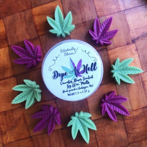 May include: A small round container of soy wax melts with a green and purple marijuana leaf design. The container has a white label with the text "Dope As Hell" and "Cannabis Flower Scented Soy Wax Melts".