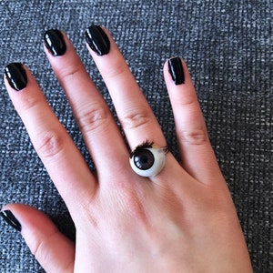 May include: A hand with black nail polish is wearing a ring with a realistic looking eyeball. The ring is gold and has a black eyelash on the top of the eyeball.