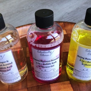 May include: Three clear plastic bottles with black lids and labels. The bottles contain pink, yellow, and clear liquid. The labels have the text "Eclectically Ellaree" and describe the scents of the liquids: "Cherry Blossom Scented 2-in-1 Bubble Bath & Shower Gel", "Going Bananas Scented 2-in-1 Bubble Bath & Shower Gel", and "Vanilla Sugar & Sandalwood Scented Bubble Bath & Shower Gel".