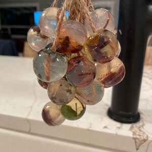 Pode incluir: Um conjunto de esferas transparentes com inclusões castanhas e verdes, suspensas por uma corda de fibra natural. As esferas, de tamanhos variados, estão agrupadas, assemelhando-se a um cacho de uvas. O fundo está desfocado.
