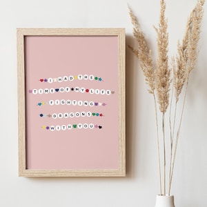 Pode incluir: Uma impressão emoldurada com as palavras "I HAD THE TIME OF MY LIFE FIGHTING DRAGONS WITH YOU" em letras coloridas de contas em um fundo rosa. A moldura é marrom claro, e a imagem está ao lado de um vaso de plantas secas.