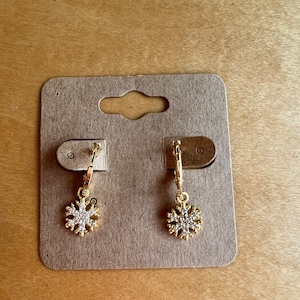 May include: A pair of gold hoop earrings with a snowflake charm. The snowflake is adorned with clear rhinestones.