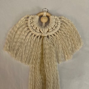 May include: A white macrame wall hanging with a wooden ring and a long tassel. The tassel is made of a cream-coloured yarn with a gold stripe.