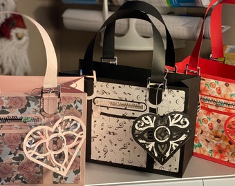 Op de afbeelding: Drie papieren tassen met verschillende ontwerpen. De eerste tas is roze met een bloemenpatroon en een hartvormige uitsparing. De tweede tas is zwart met een notenpatroon en een zwarte hartvormige uitsparing. De derde tas is rood met een bloemenpatroon en een rode hartvormige uitsparing.