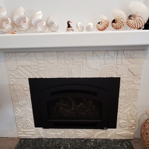 May include: A white tiled fireplace with a black fireplace insert. The fireplace is decorated with white ceramic tiles in a geometric pattern. There are several nautilus shells on the mantel above the fireplace.