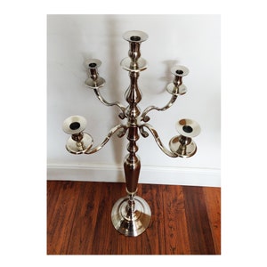 May include: A polished silver candelabra with five candle holders. The candelabra features a decorative, tall base and curved arms. Each candle holder is circular with a dark interior. The metal candelabra has a shiny finish and is suitable for various settings.