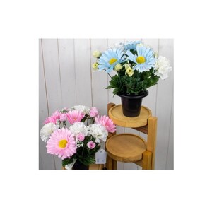 May include: Two bouquets of artificial flowers in black pots. One bouquet features pink and white daisies, the other has blue daisies. The flowers are displayed on a wooden tiered plant stand against a white wood-panelled background.