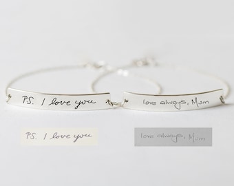 Puede incluir: Dos pulseras de plata con un mensaje escrito a mano en cada una. La primera pulsera dice "P.S. Te quiero" y la segunda pulsera dice "Te quiero siempre, Mamá".
