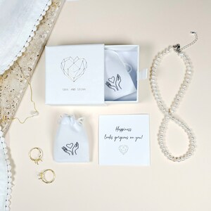 Puede incluir: Una caja de regalo blanca con un dise&ntilde;o de coraz&oacute;n geom&eacute;trico y el texto "Soul and Silva". La caja contiene una bolsa blanca con un dise&ntilde;o de coraz&oacute;n y manos. Tambi&eacute;n se muestra una tarjeta blanca con el texto "Happiness looks gorgeous on you!" y un collar de perlas.