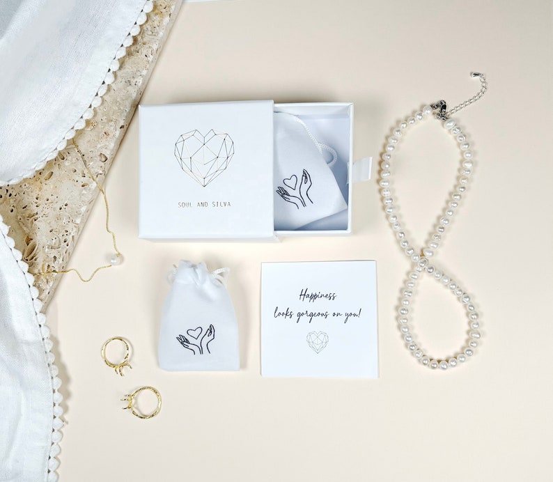 Puede incluir: Una caja de regalo blanca con un dise&ntilde;o de coraz&oacute;n geom&eacute;trico y el texto "Soul and Silva". La caja contiene una bolsa blanca con un dise&ntilde;o de coraz&oacute;n y manos. Tambi&eacute;n se muestra una tarjeta blanca con el texto "Happiness looks gorgeous on you!" y un collar de perlas.