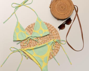 Könnte beinhalten: Ein zweiteiliger Bikini mit hellgrünem und gelbem Polka-Dot-Muster. Das Bikini-Oberteil ist im Neckholder-Stil gehalten, und die Hose ist zum Binden. Eine runde gewebte Tasche, Sonnenbrille und ein gewebter Platzdeckchen sind ebenfalls im Bild.