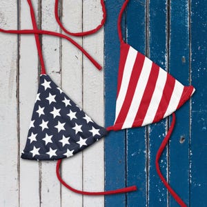 May include: A patriotic bikini top featuring the American flag design. One cup displays a navy blue background with white stars, while the other has red and white stripes. The straps are red, and the background is a weathered wood.