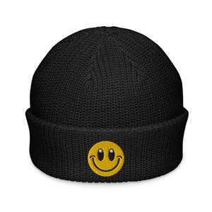 May include: Black knit beanie with a yellow embroidered smiley face. The beanie has a folded cuff and a ribbed texture. The smiley face has black eyes and a curved smile.