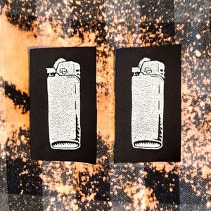 Puede incluir: Dos parches de tela negra con imágenes bordadas blancas de encendedores.