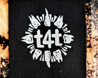 Peut inclure: Patch en tissu noir avec un motif brodé blanc. Le motif représente un cercle avec le texte "t4t" au centre et une série de dents stylisées autour du cercle.