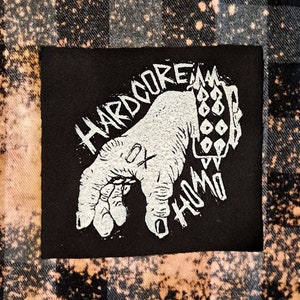 Könnte beinhalten: Ein schwarz-weißes Patch mit dem Text "HARDCORE HOMO" und einer Hand mit einem Armband. Das Patch ist auf einen schwarz-weiß karierten Stoff genäht.