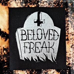 May include: Black and white iron-on patch with a tombstone design. The tombstone has a cross on top and the words "BELOVED FREAK" written on it.