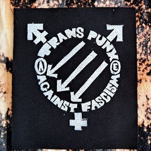 Può includere: Toppa in tessuto nero con un cerchio bianco che contiene tre linee diagonali e il testo "Trans Punk Against Fascism" attorno al cerchio. Il cerchio è affiancato da un simbolo transgender a sinistra e da una freccia che punta verso l'alto a destra.
