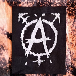 Könnte beinhalten: Schwarzer Stoff-Patch mit einem weißen Anarchiesymbol, einem stilisierten "A" in einem Kreis mit strahlenden Linien.