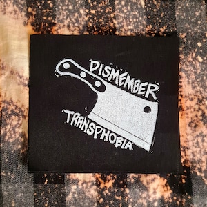 Op de afbeelding: Zwarte stoffen patch met witte tekst en een witte illustratie van een slagersmes. De tekst luidt "DISMEMBER TRANSPHOBIA".