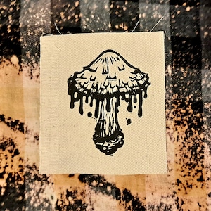 Op de afbeelding: Een zwart-witte illustratie van een paddenstoel met druppelende randen, gedrukt op een beige stoffen patch.