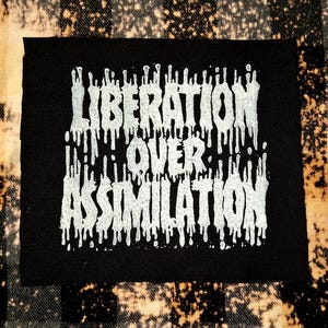 Könnte beinhalten: Schwarzer quadratischer Aufnäher mit dem Schriftzug "LIBERATION OVER ASSIMILATION" in tropfenden weißen Buchstaben. Der Aufnäher ist vor einem karierten Hintergrund mit gebleichten Akzenten platziert.