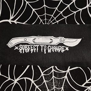 Könnte beinhalten: Schwarzer Patch mit einer weißen Umrandung eines Skalpells und dem Text "Subject to Change" in Weiß.