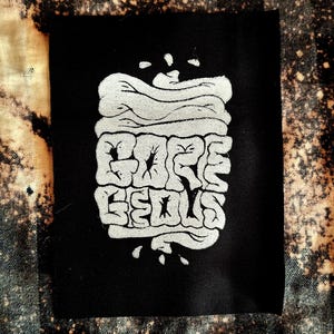 Op de afbeelding: Zwarte patch met een witte afbeelding van een gestileerde, gelaagde, organische vorm met de tekst "Gore Geous" erin.