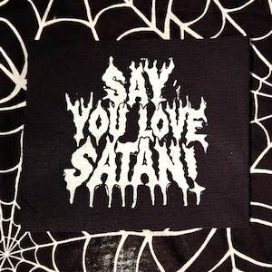 May include: A rectangular black fabric patch featuring white screen-printed text that reads SAY YOU LOVE SATAN! in a stylized, dripping, jagged font. The lettering is centered on the patch, which has a flat, matte finish and clean edges.