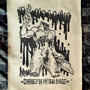 Puede incluir: Parche en blanco y negro que representa una figura sin cabeza sosteniendo una cuchilla y una sierra, con gotas negras. El texto "CARVED IN MY OWN IMAGE" está en la parte inferior del parche.