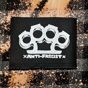 May include: Black patch with a white image of a knuckle duster and the text "xANTI-FASCISTx" printed below.